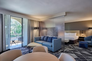 Hotellihuoneessa on sohva, sänky, pöytä ja tuoli. Huoneesta on parveke, jolta avautuu näkymä puita kohti.