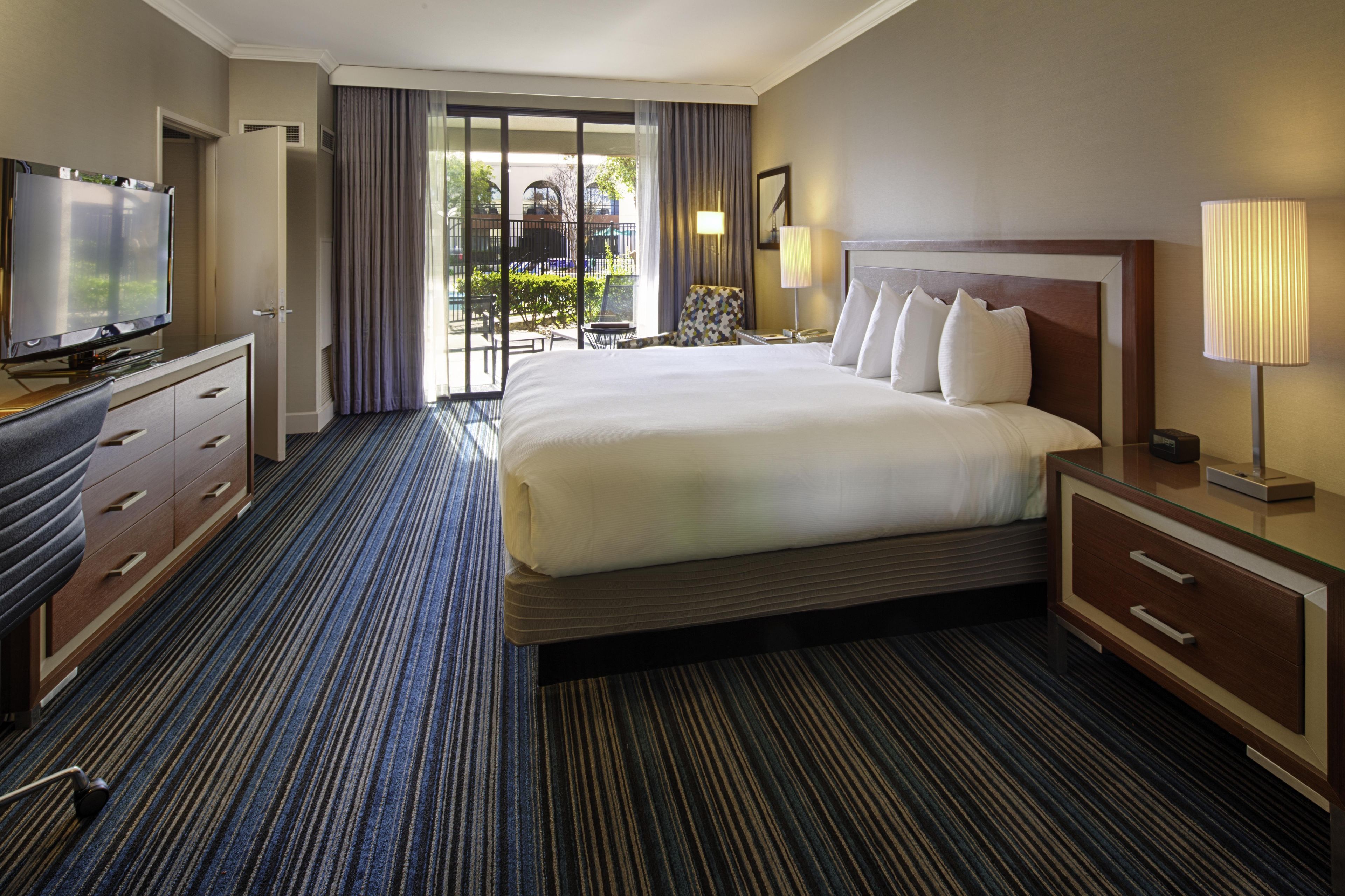 Habitación de hotel con una cama grande, televisor, escritorio con cajones y vistas al exterior a través de una puerta corredera.