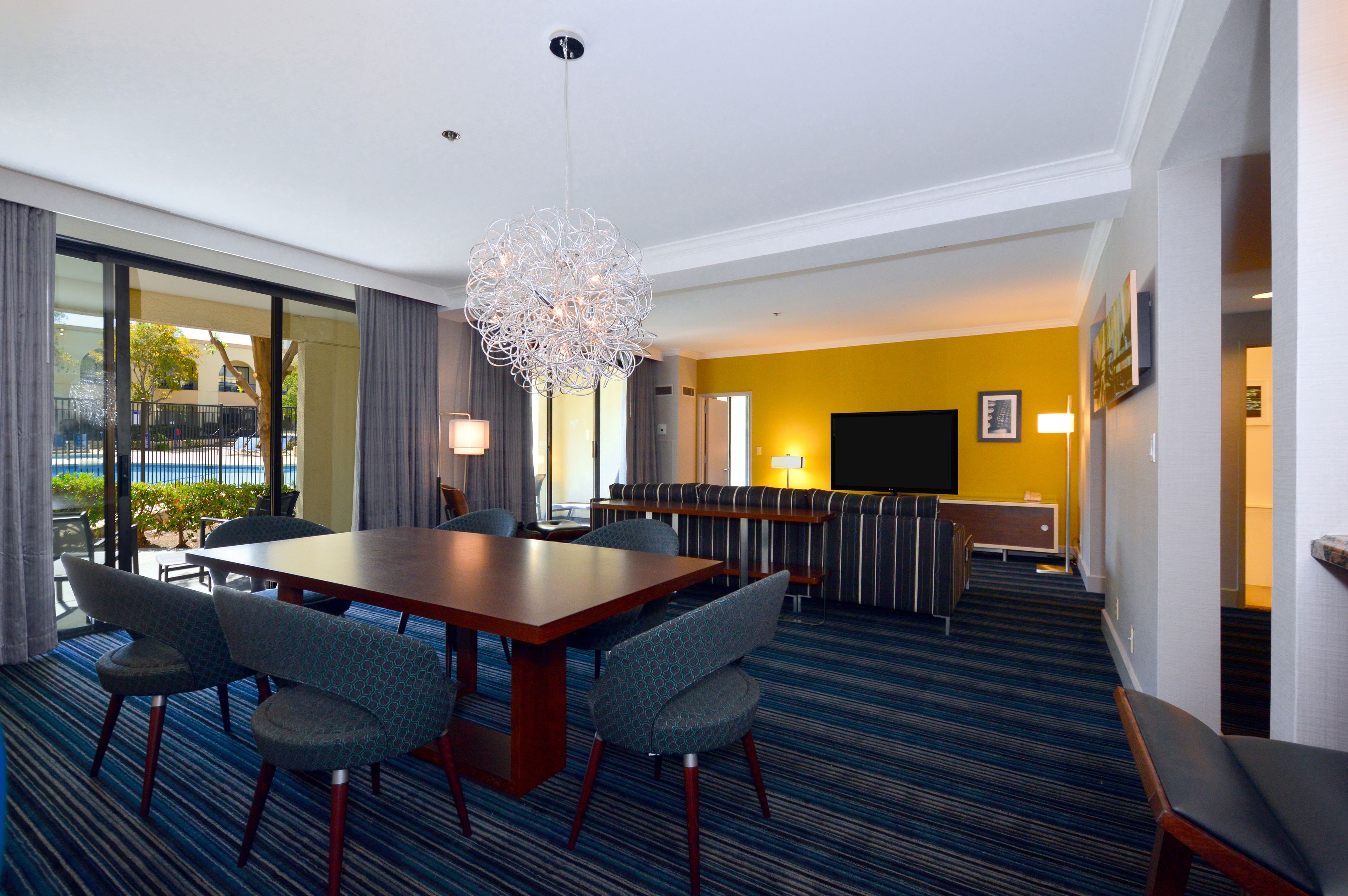 A hotel room with a large dining table, comfortable seating, a sofa, a TV, and a view of the outdoors.