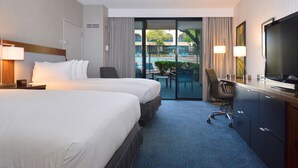 A hotel room with two beds, a desk, a TV, and a view of a pool and outdoor area. - Doubletree by Hilton Newark - Fremont (Newark)
