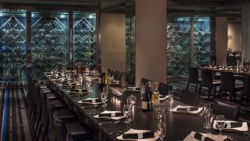 Uma longa mesa de jantar posta para uma refeição, com garrafas de vinho, copos e guardanapos dobrados com cuidado.