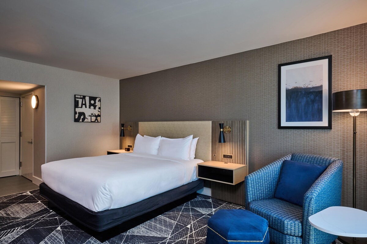 A hotel room with a large bed, a nightstand, a chair, and a small table.
