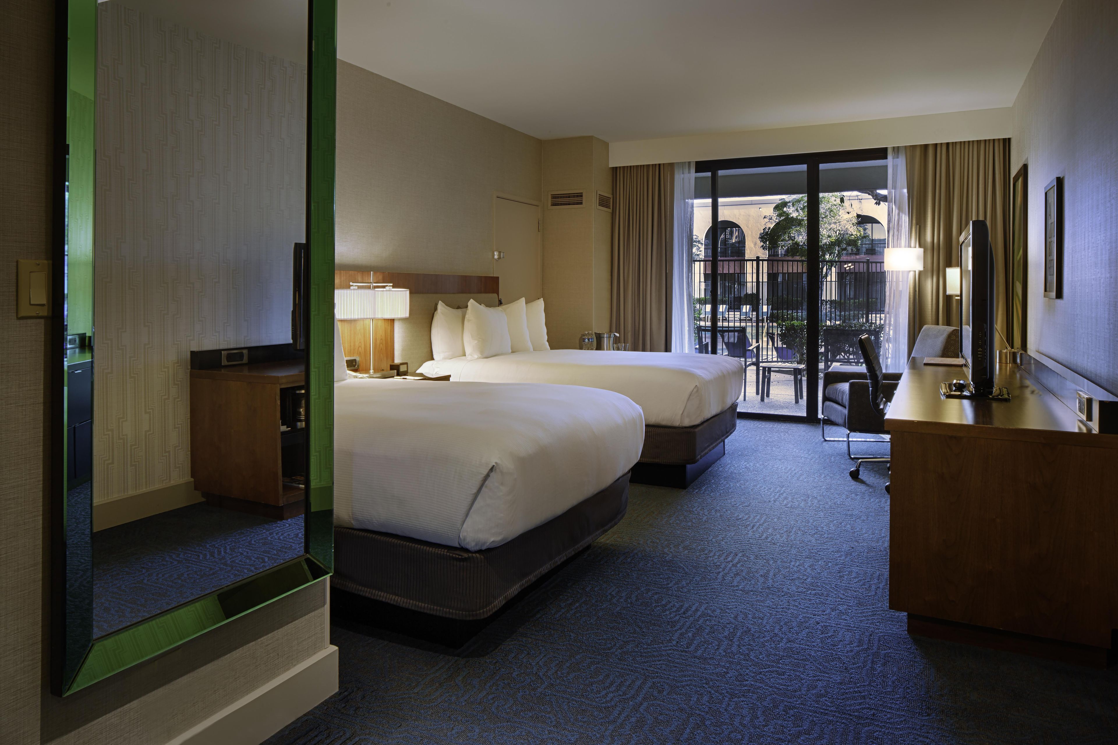 A hotel room with two beds, a desk, a TV, and a balcony with a view.