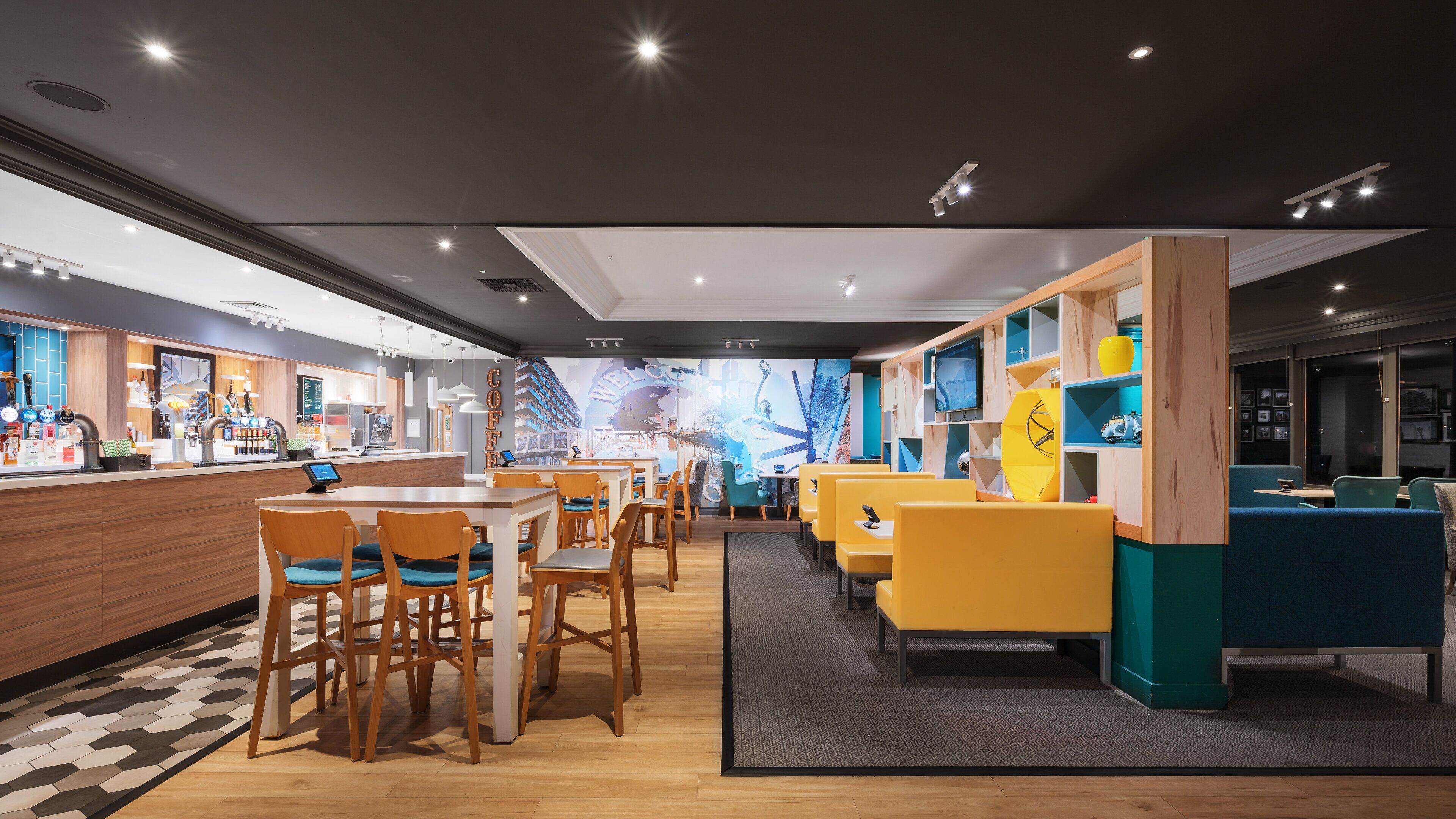 Un café moderne avec des tables et des chaises en bois, un bar doté de robinets et une fresque colorée sur le mur.