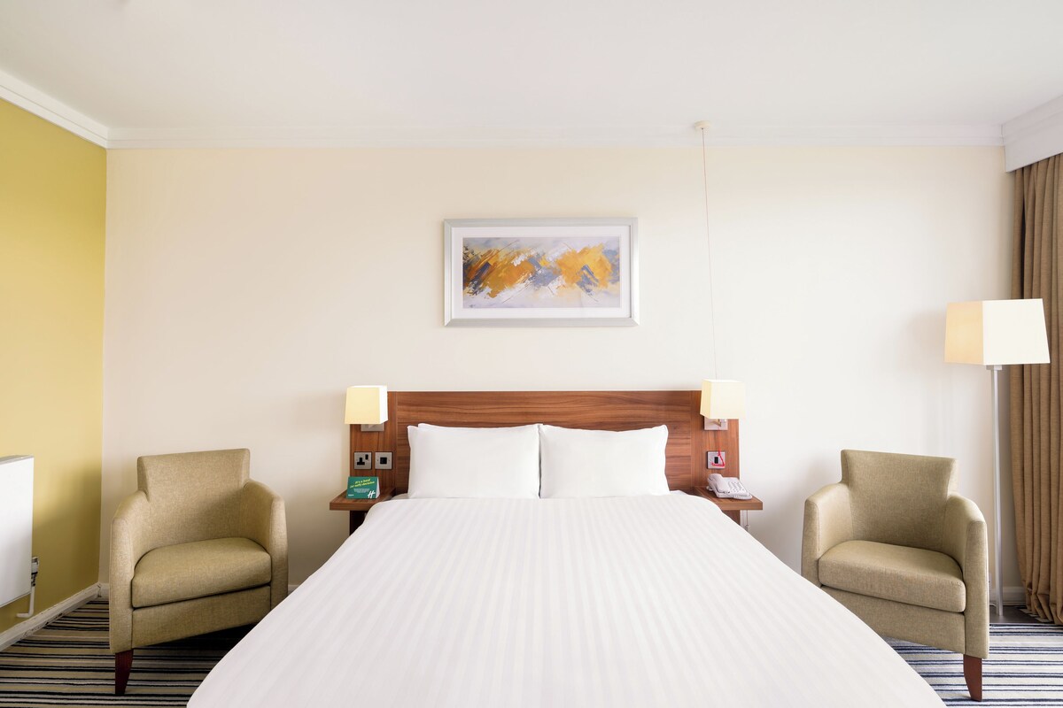 A hotel room with a large bed, two armchairs, a wooden headboard, a painting on the wall, and a lamp on the nightstand.