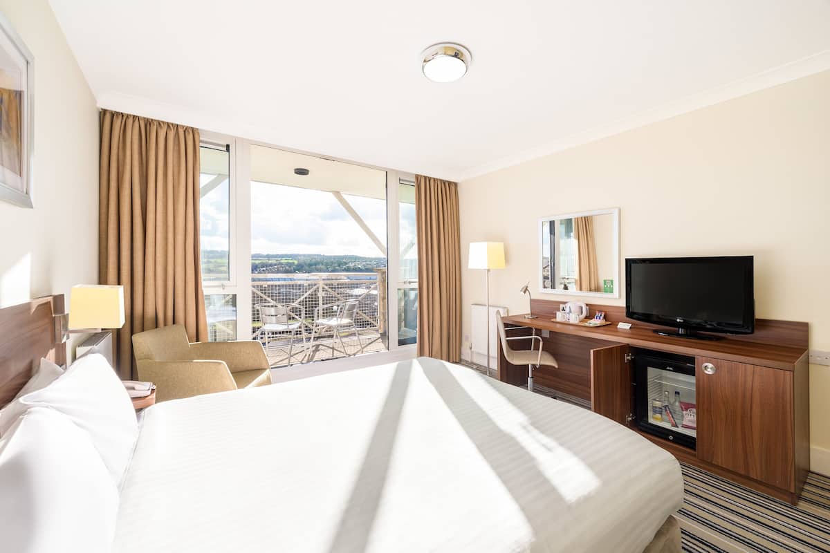 A hotel room with a large bed, a desk with a television, a chair, and a view of the city through large windows.