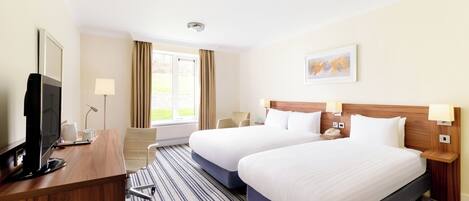 Habitación de hotel con dos camas, un escritorio con computadora, una silla, una ventana con cortinas y un cuadro en la pared.
