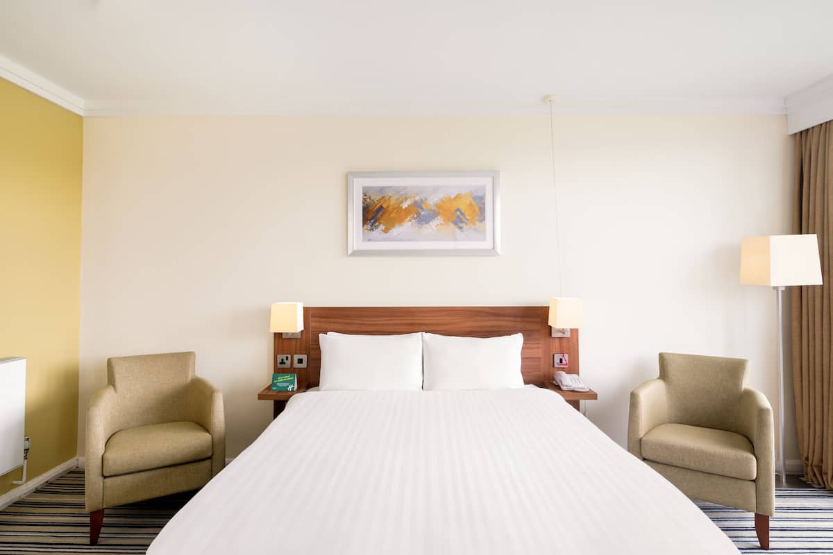A hotel room with a large bed, two armchairs, a wooden headboard, a painting on the wall, and a lamp on the nightstand.