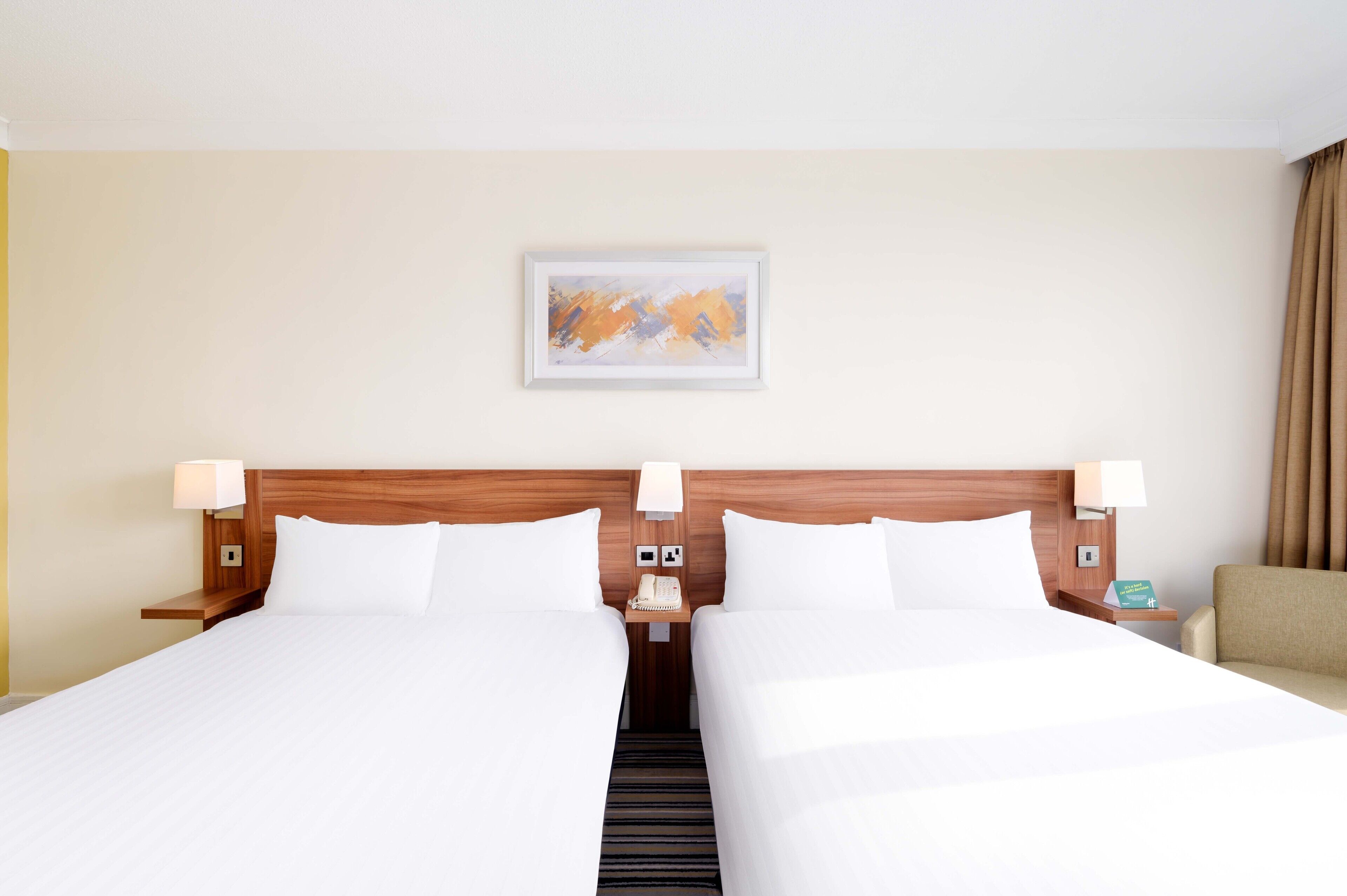 A hotel room with two beds, a wooden headboard, bedside lamps, a nightstand with a phone, and a painting on the wall.