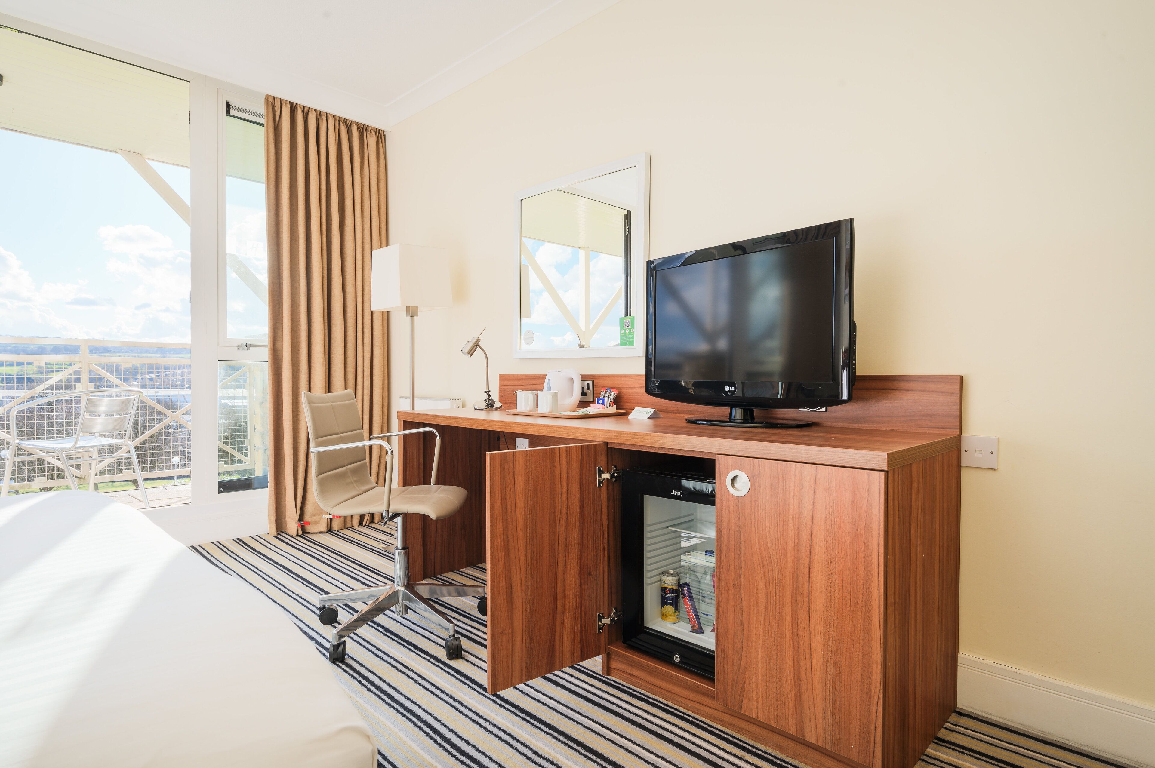 Une chambre d’hôtel dotée d’une grande fenêtre, d’un bureau avec une télévision, d’une chaise et d’un mini-bar.