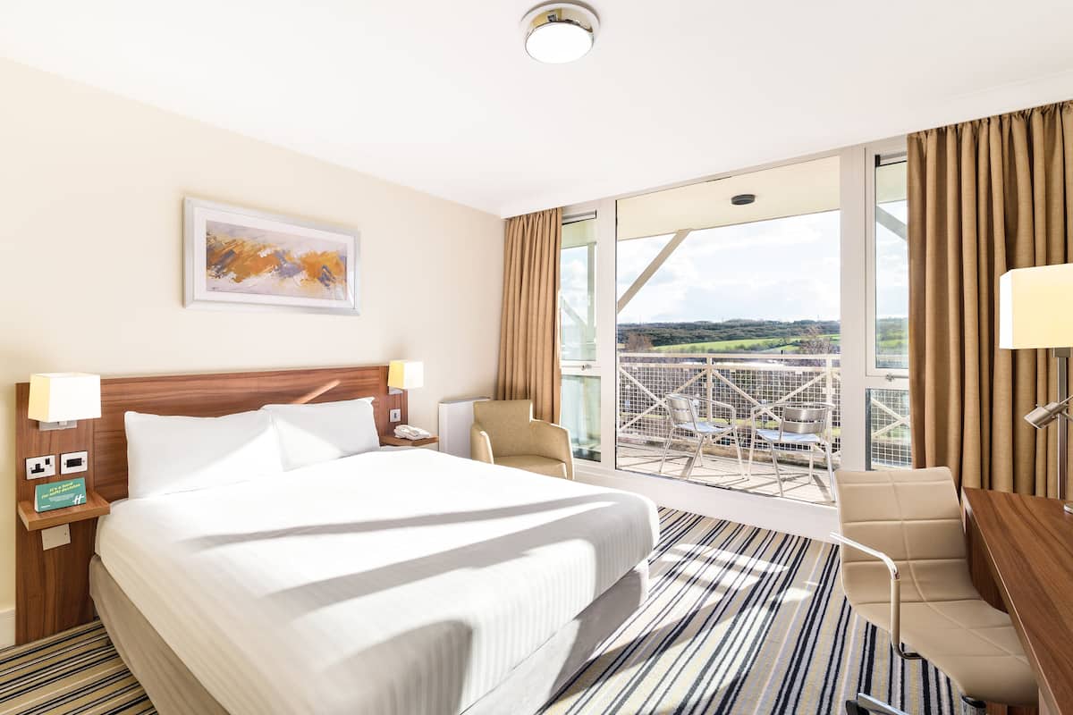 A hotel room with a large bed, a desk, a chair, a balcony with a view, and a painting on the wall.