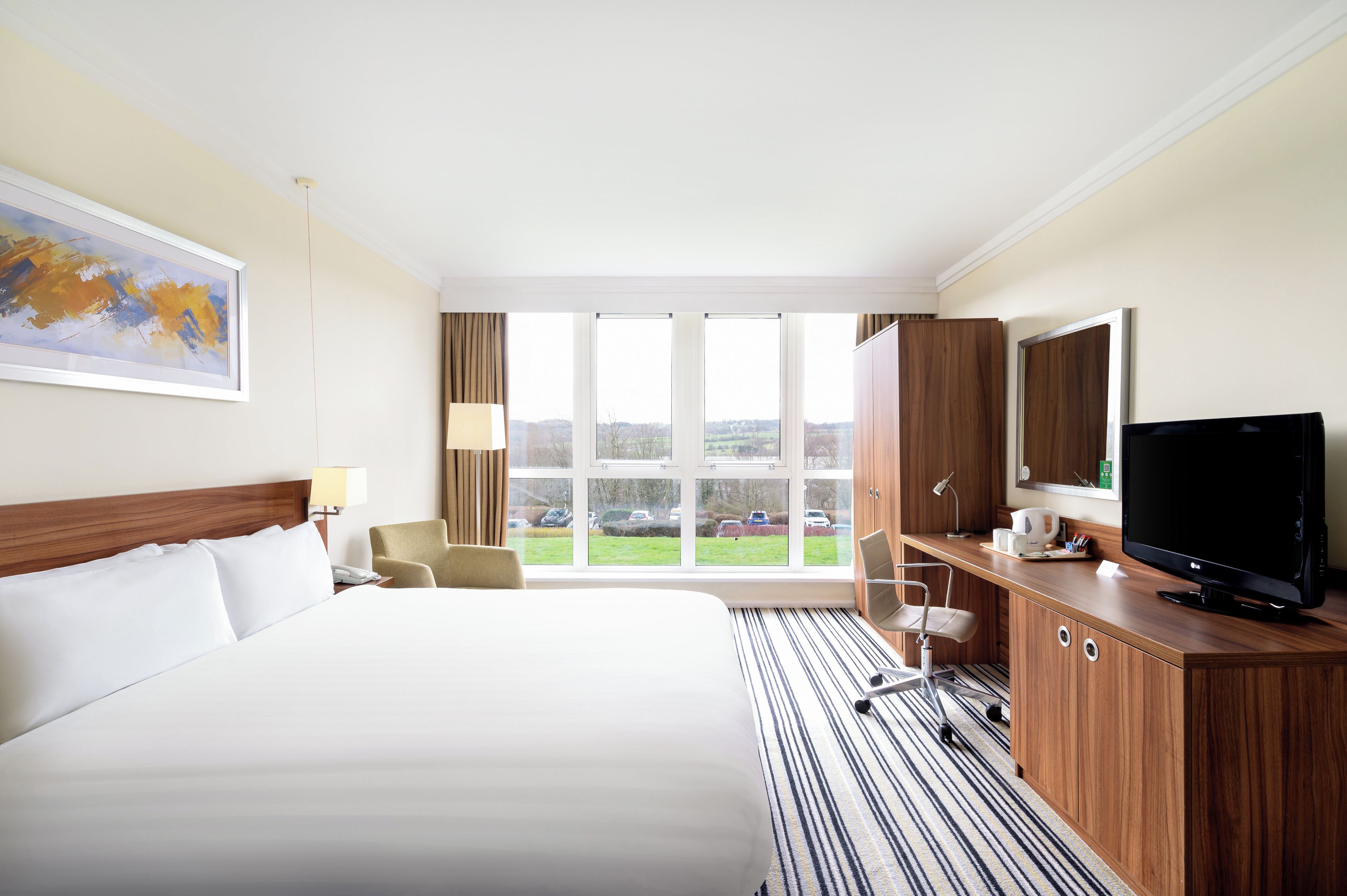 A hotel room with a large bed, a desk with a TV, a chair, a window with a view of the parking lot, and a painting on the wall.