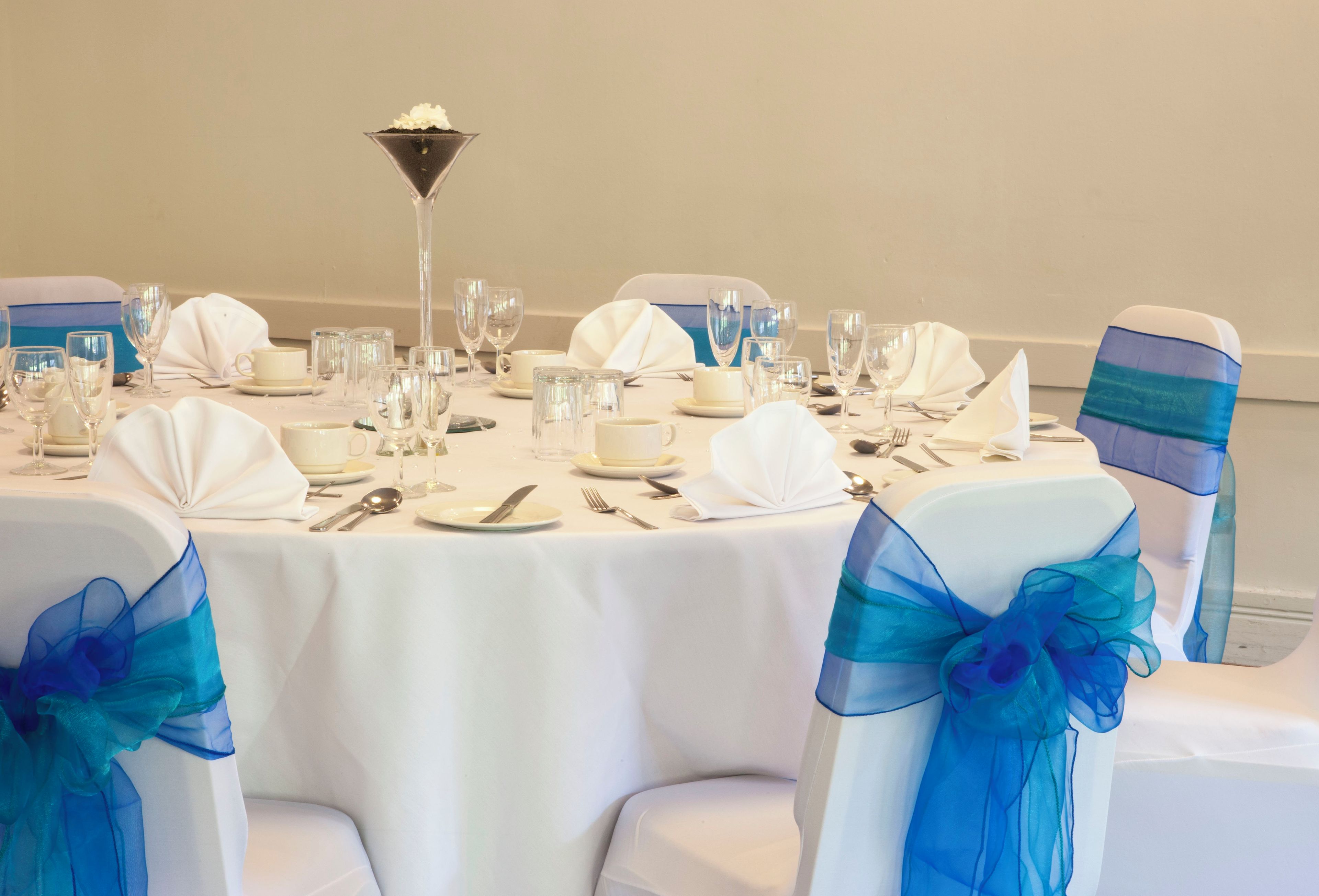 Een ronde tafel gedekt voor een formeel evenement, met witte tafellakens, glazen servies en blauwe stoellinten.