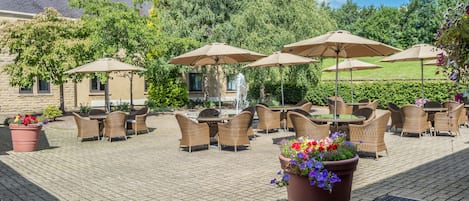 An outdoor patio with wicker furniture and umbrellas, surrounded by greenery and a building in the background.