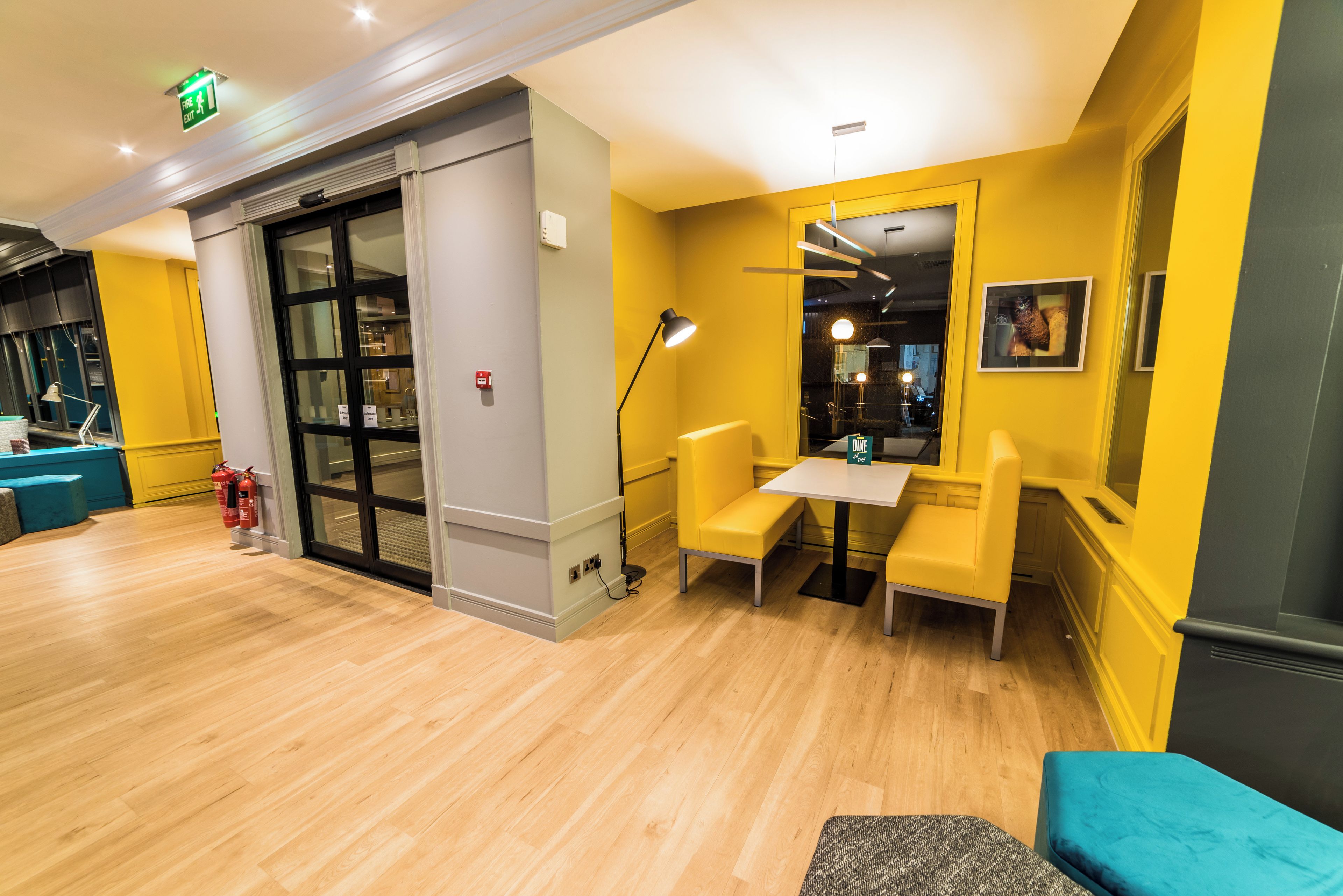 A reception area with a wooden floor, a blue sofa, a table with chairs, a fire exit sign, and a fire extinguisher.
