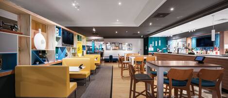 A modern restaurant with yellow and blue seating, wooden accents, and a bar area.