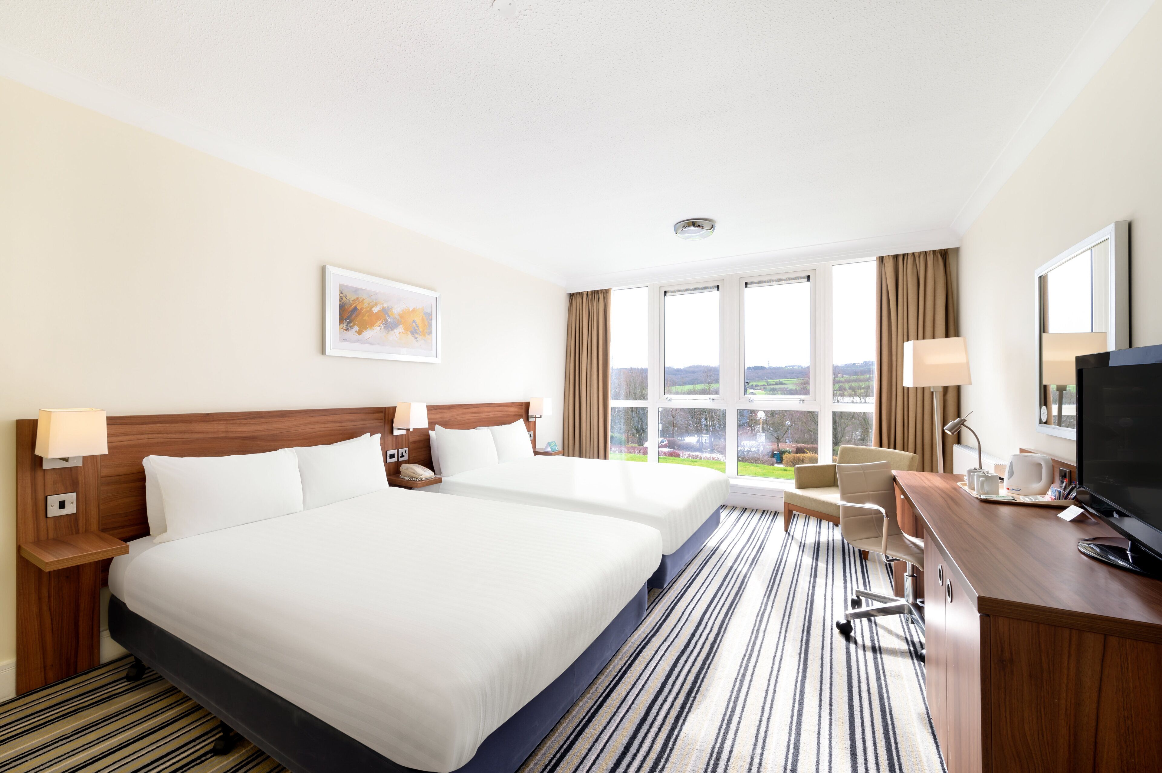 A hotel room with two beds, a desk, a chair, a TV, and large windows offering a view of the outdoors.