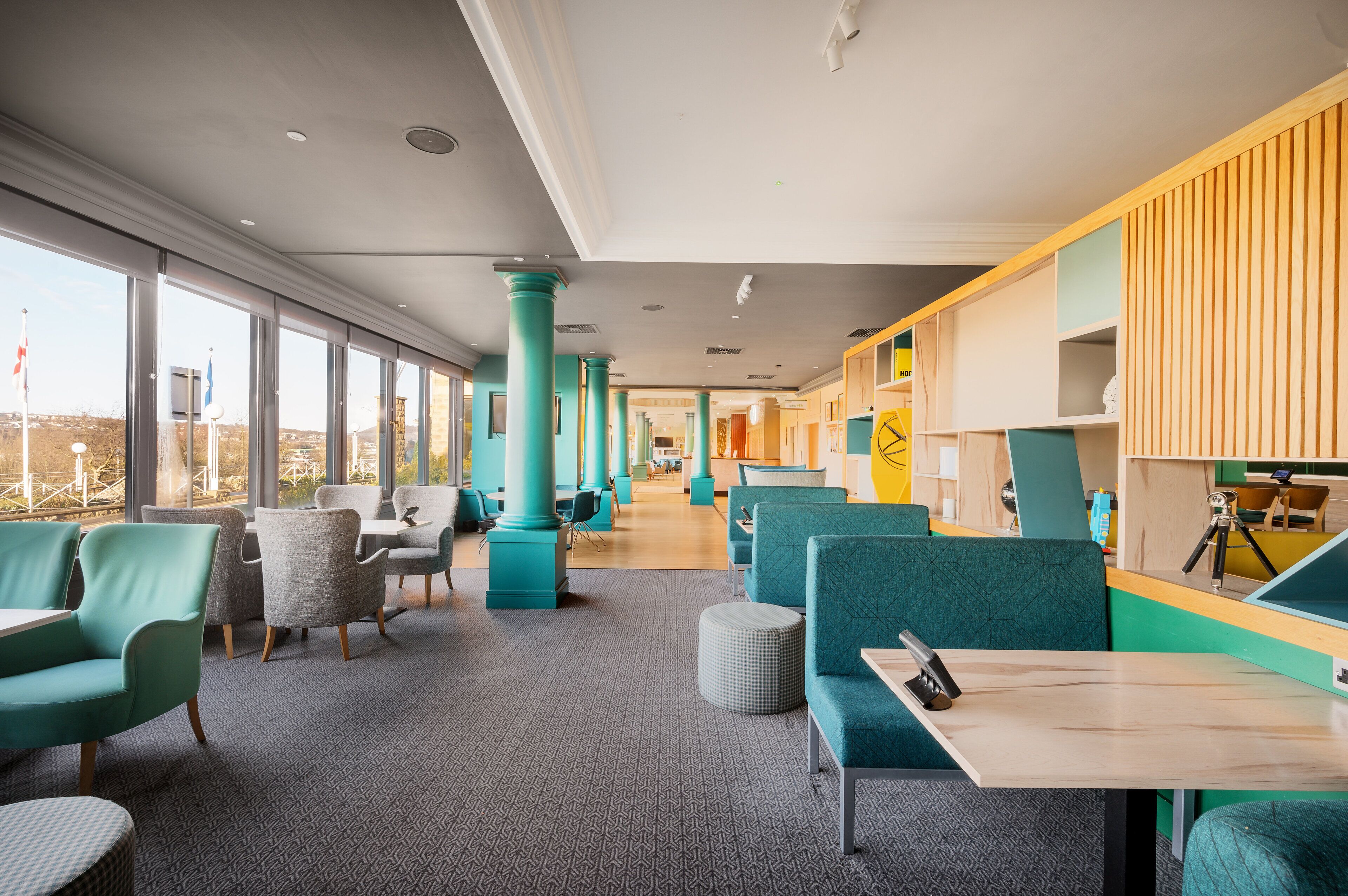 A modern interior with teal and gray seating, large windows, and a bright yellow accent wall.