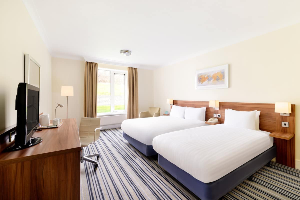 A hotel room with two beds, a desk with a computer, a chair, a window with curtains, and a painting on the wall.