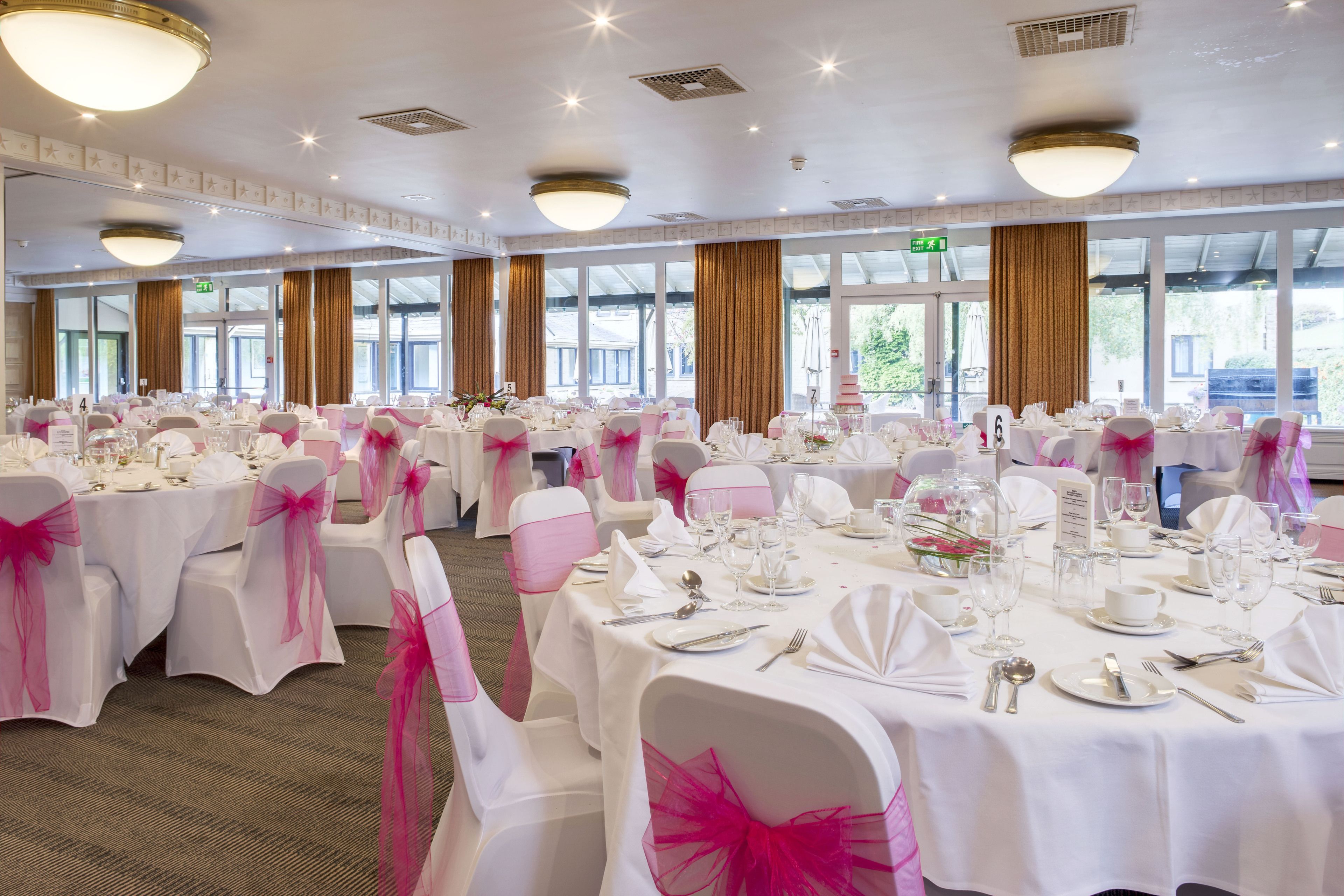 Un salón de banquetes con mesas redondas dispuestas para cenar, manteles blancos y cintas rosas en las sillas.