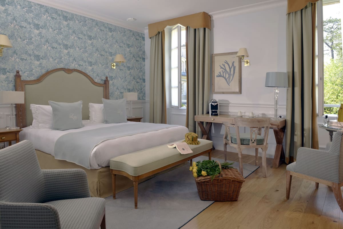 Habitación de hotel con una cama grande, un escritorio, dos sillas, un banco y una cesta de mimbre con flores.