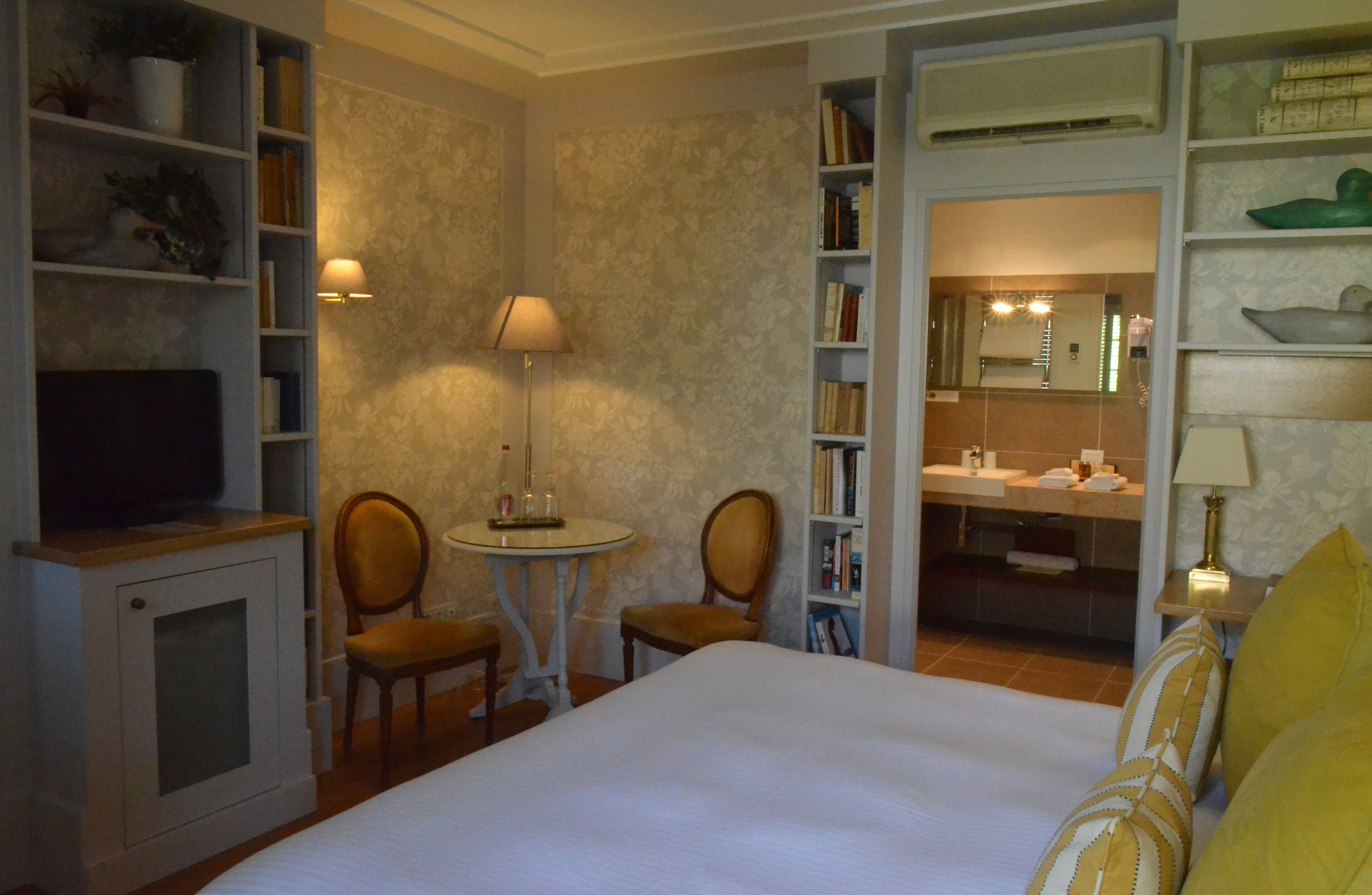 A hotel room with a bed, a desk with a lamp, chairs, a bookshelf, and a bathroom visible through an open door.