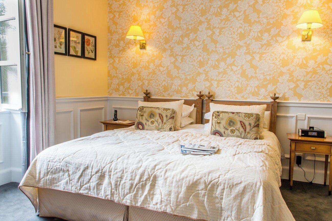 A neatly made bed with a quilt, two bedside tables with lamps, and a floral wallpaper pattern.