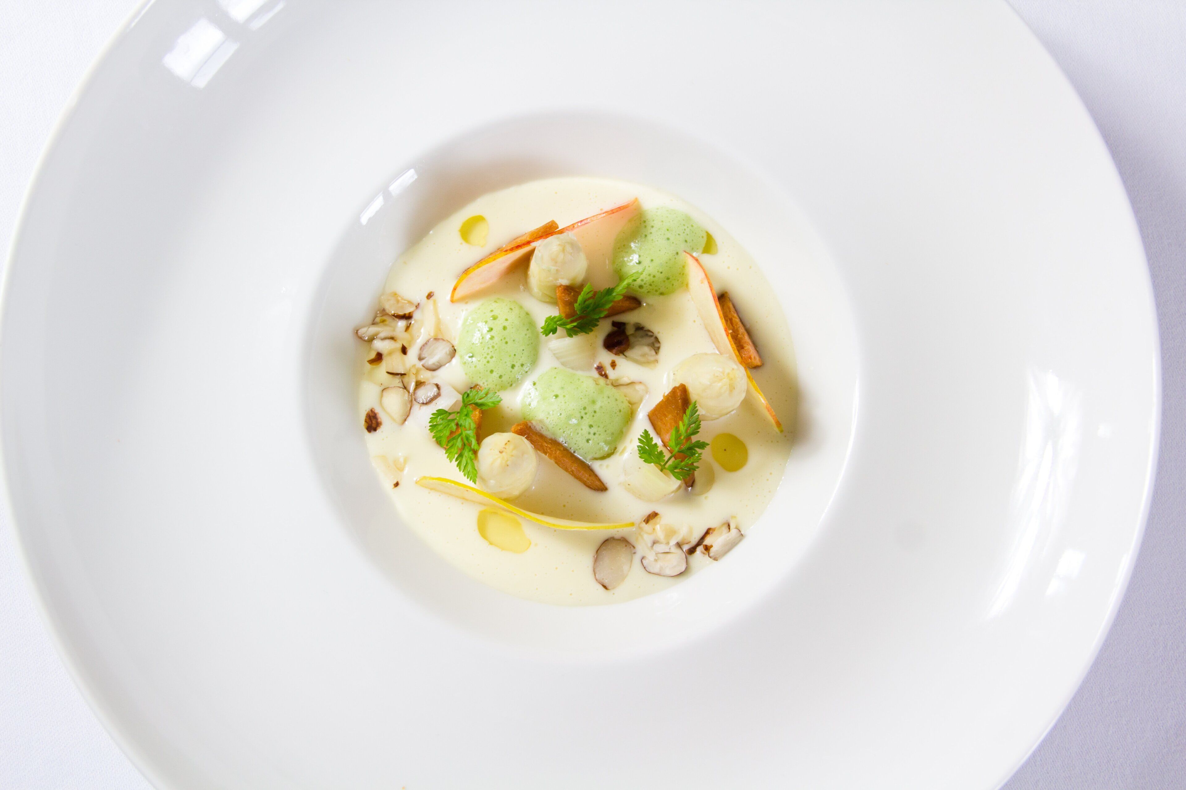 A white plate with a creamy dessert featuring green spheres, nuts, and a garnish.