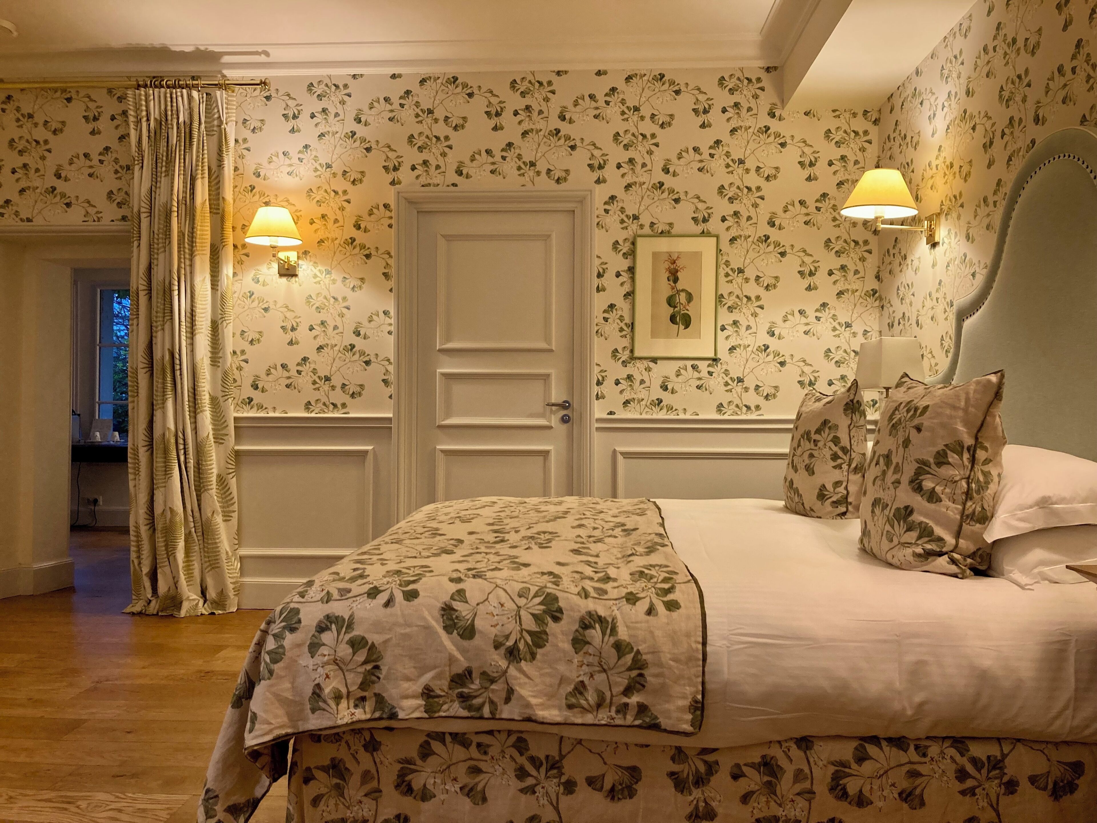 Ein Schlafzimmer mit floraler Tapete, einem Bett mit passenden Kissen, zwei Wandleuchten und einer Tür.