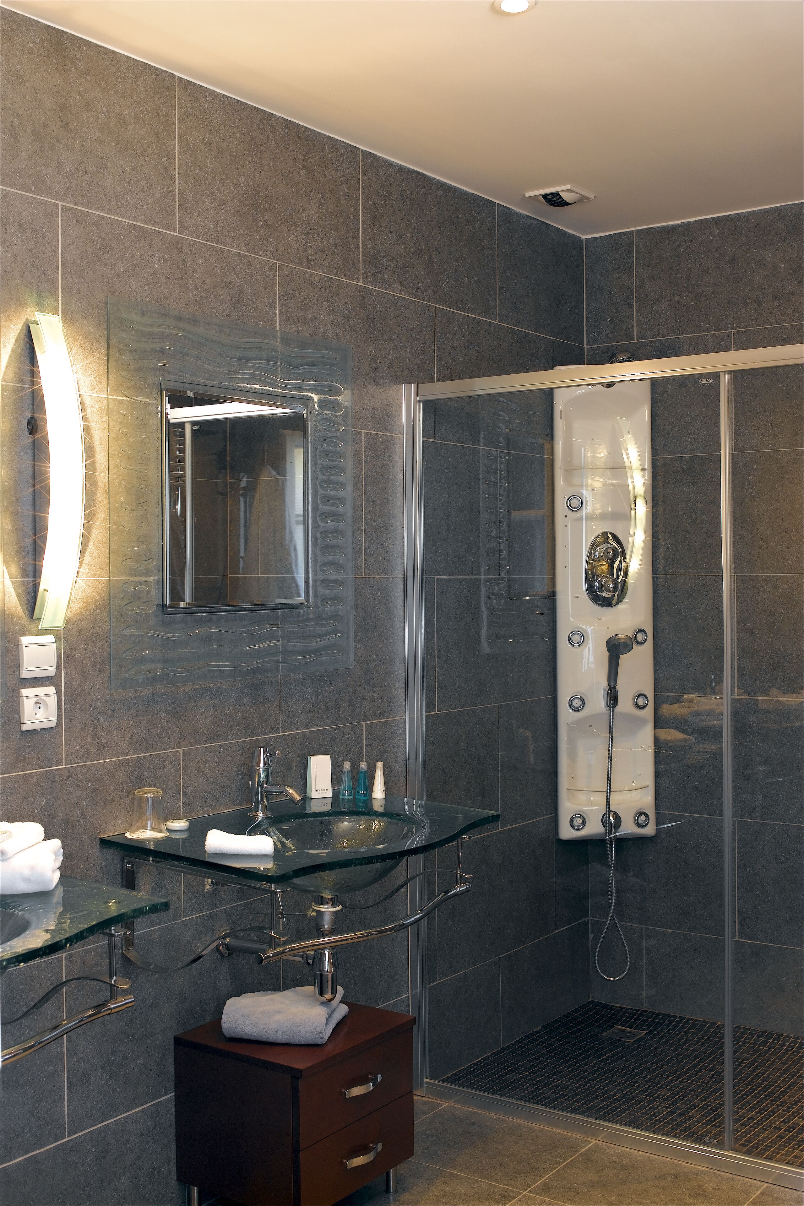 Un baño moderno con cabina de ducha de vidrio, lavamanos de cristal y tocador de tonos oscuros.