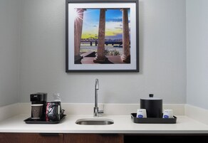 Una cocina con cafetera, pileta y un cuadro de un puente.