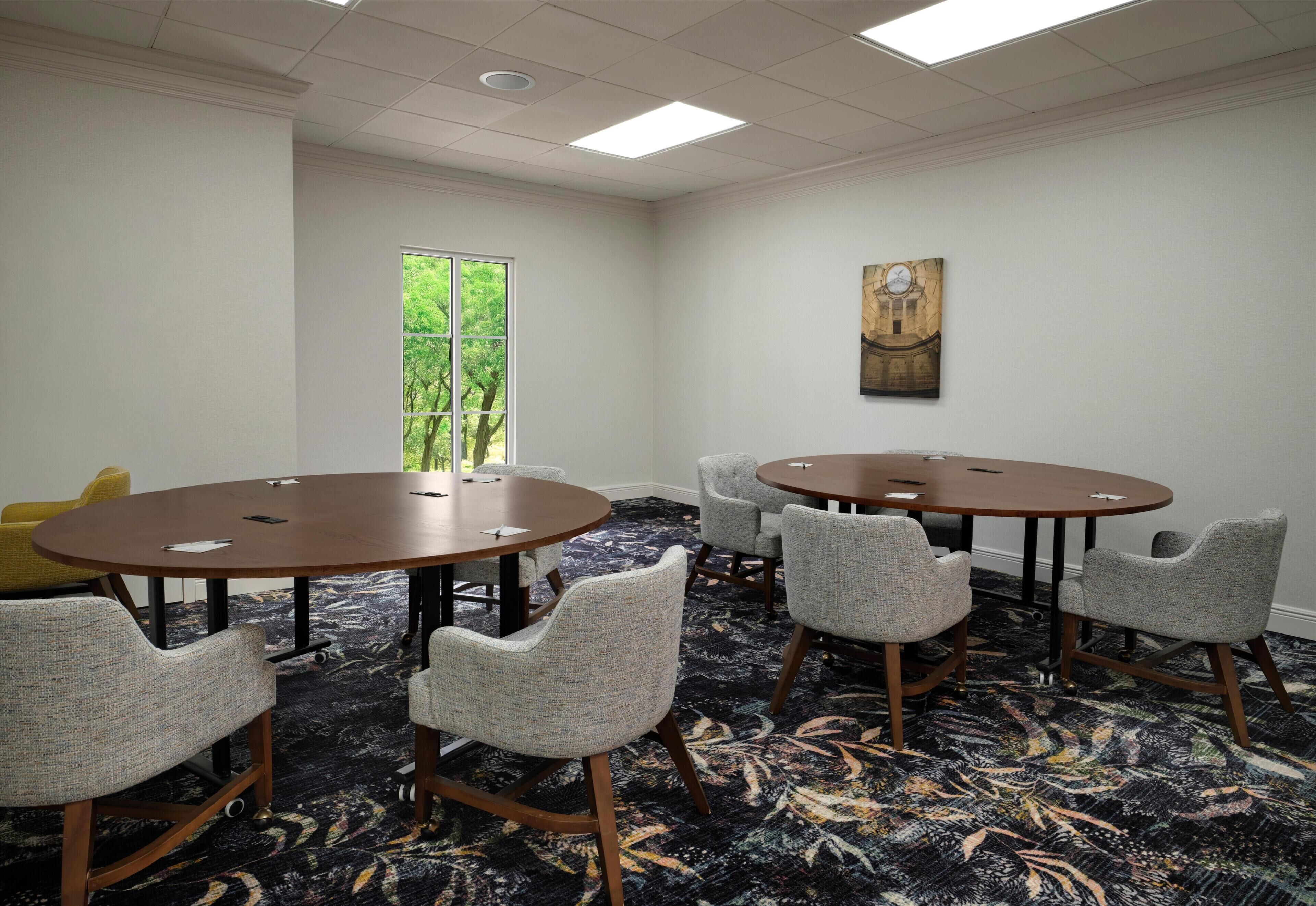 A conference room with two round tables, six upholstered chairs, a large window with a view of trees, and a painting on the wall.