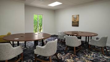 A conference room with two round tables, six upholstered chairs, a large window with a view of trees, and a painting on the wall.