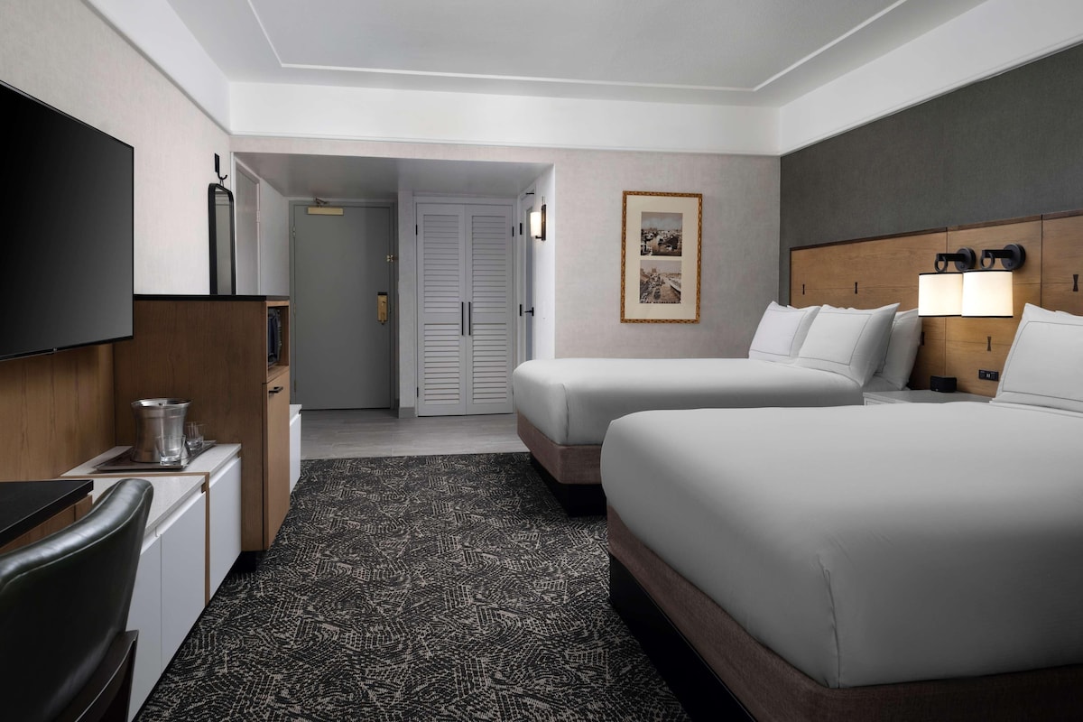 Habitación de hotel con dos camas, televisión, minibar y puerta que da a un pasillo.
