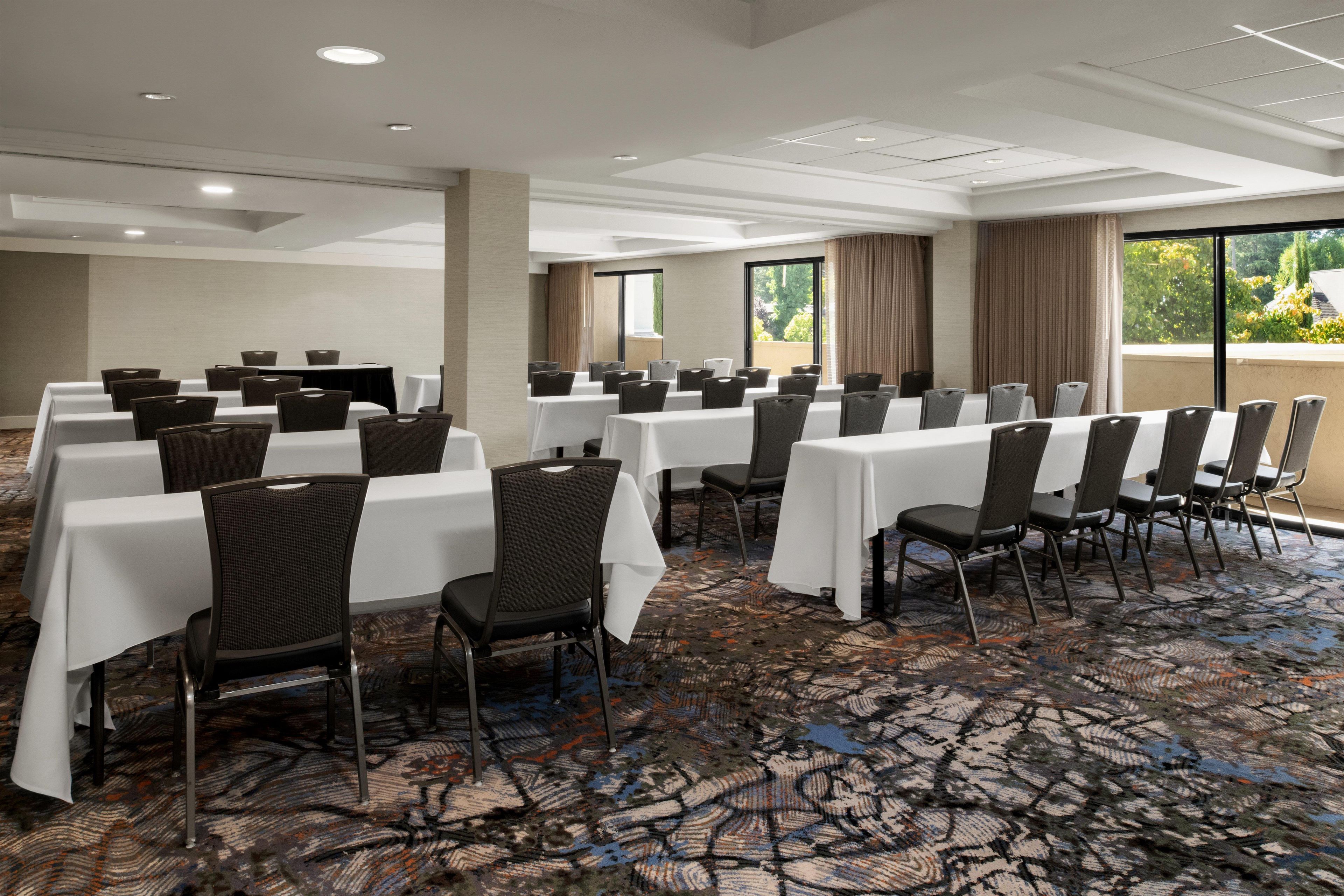 A conference room with multiple tables and chairs, large windows with curtains, and a patterned carpet.