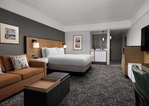 Habitación de hotel con cama, sofá, escritorio, televisión y baño visible a través de una puerta abierta.