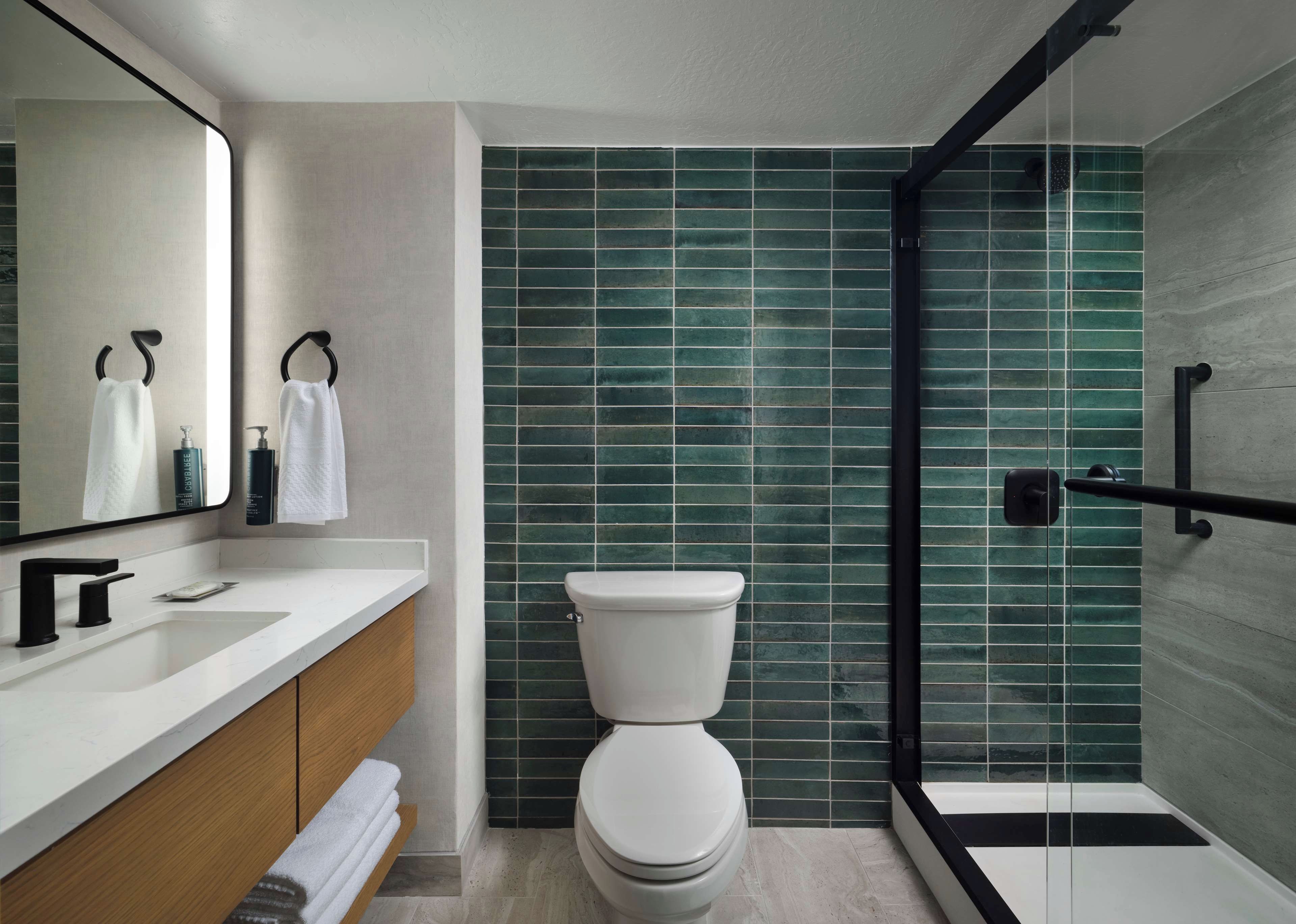 Un baño moderno con inodoro blanco, tocador de madera, espejo grande y zona de ducha con azulejos verdes.