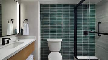 Un baño moderno con inodoro blanco, tocador de madera, espejo grande y zona de ducha con azulejos verdes.