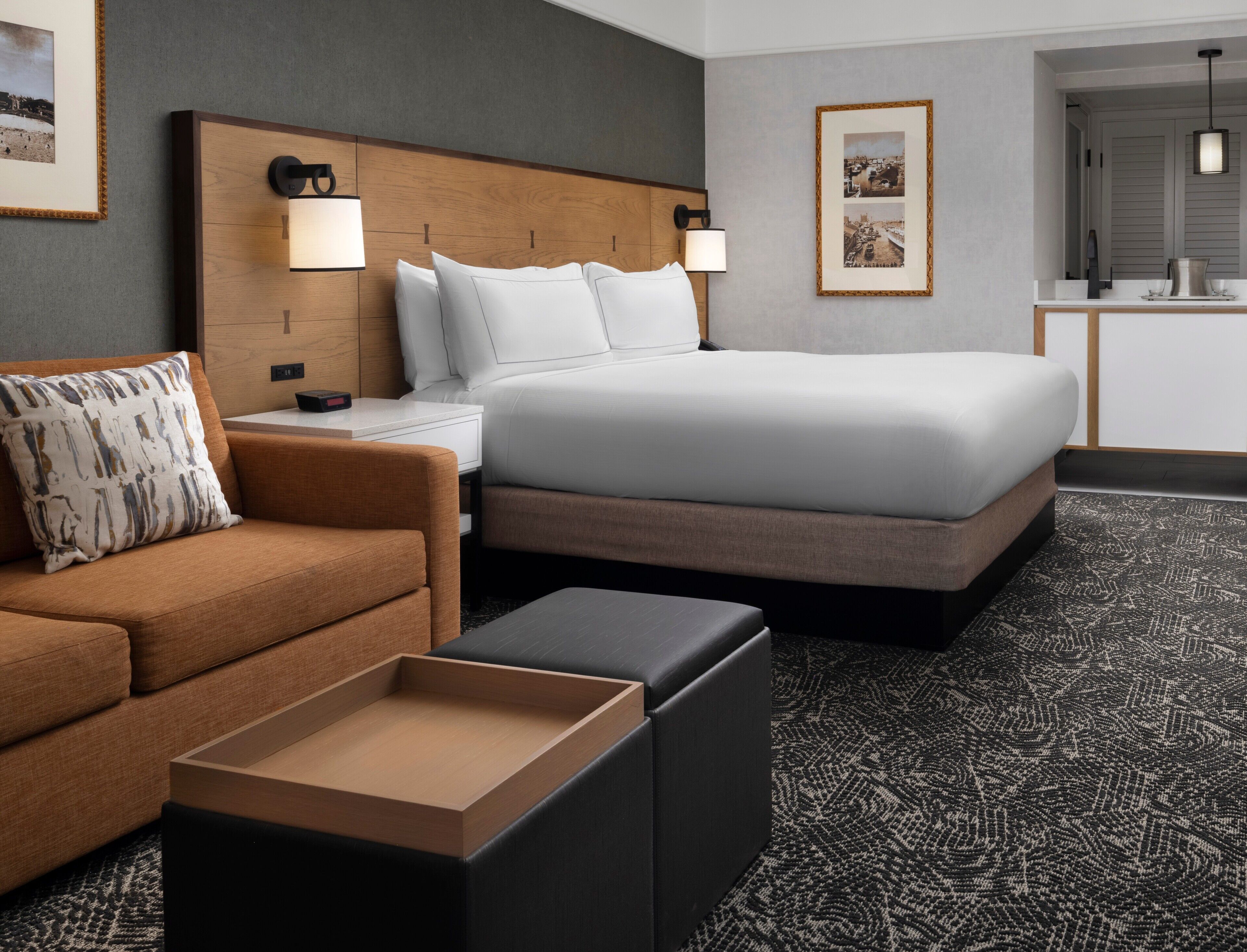 Habitación de hotel con cama, sofá, mesita de noche, lámpara, un cuadro en la pared y alfombra.