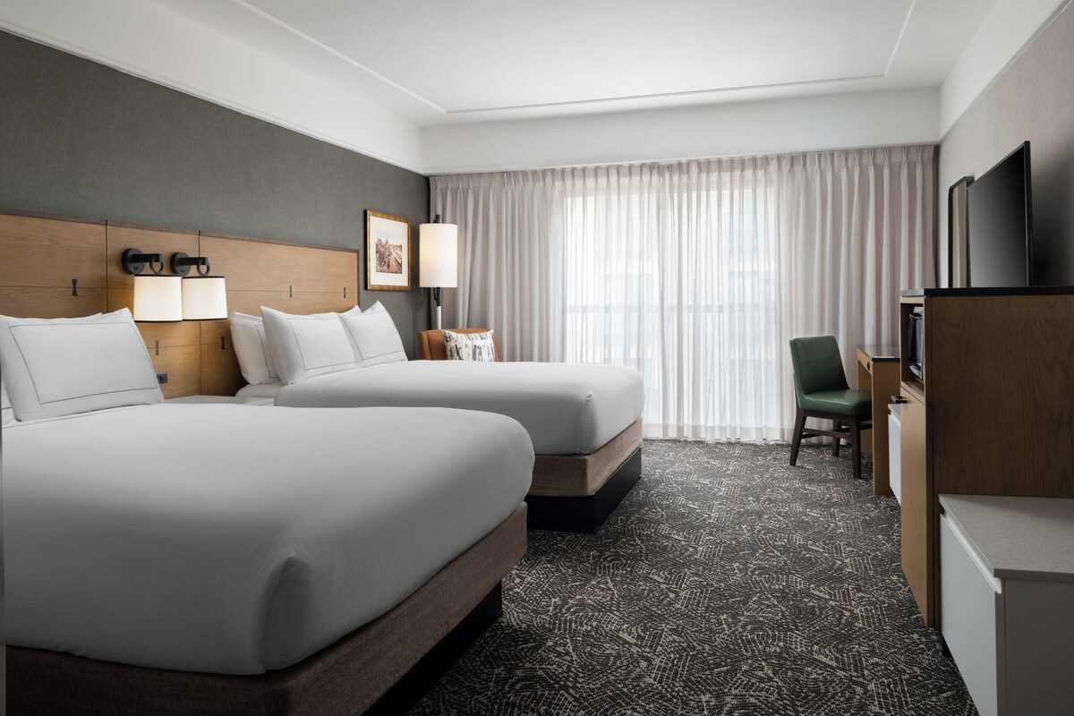 A hotel room with two beds, a desk, a chair, a TV, and a large window with curtains.