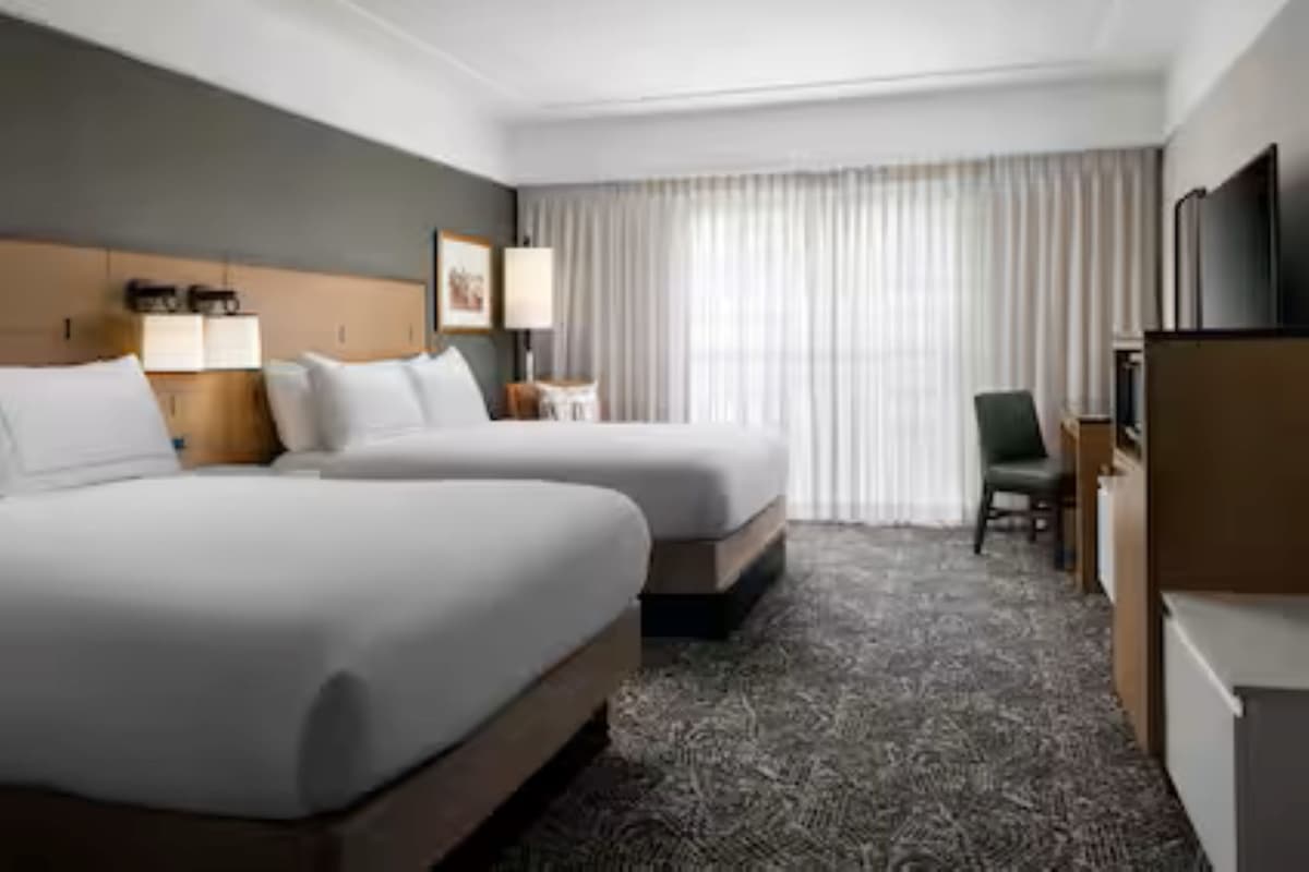 Habitación de hotel con dos camas, un escritorio, una silla, televisión y un ventanal con cortinas.