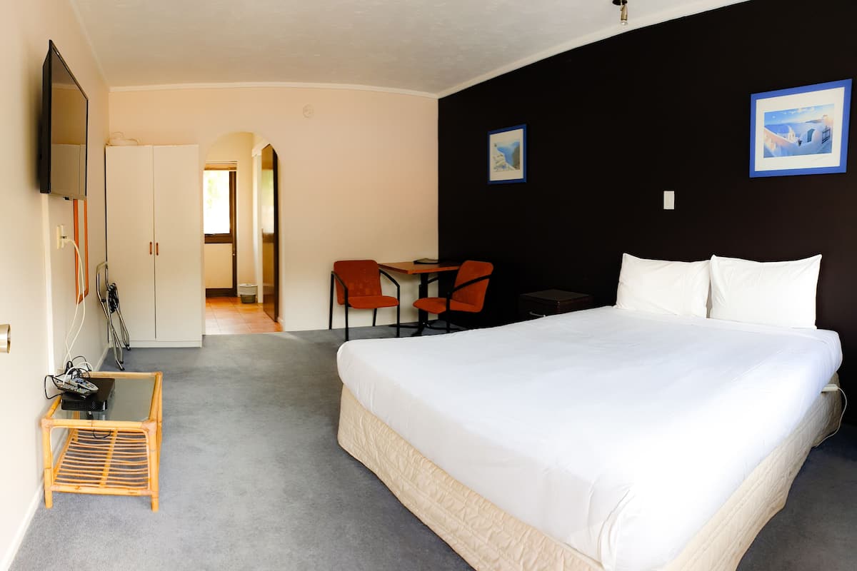Ein Hotelzimmer mit einem großen Bett, einem Schreibtisch mit zwei Stühlen, einem kleinen Tisch, einem an der Wand montierten Fernseher und zwei gerahmten Bildern.
