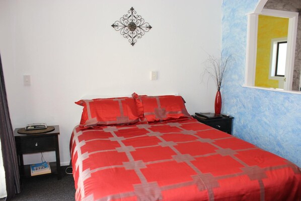 A bedroom with a red bedspread, a nightstand with a radio, a wall decoration, and a window with a yellow frame. - Casa Bella Motel (Paihia)