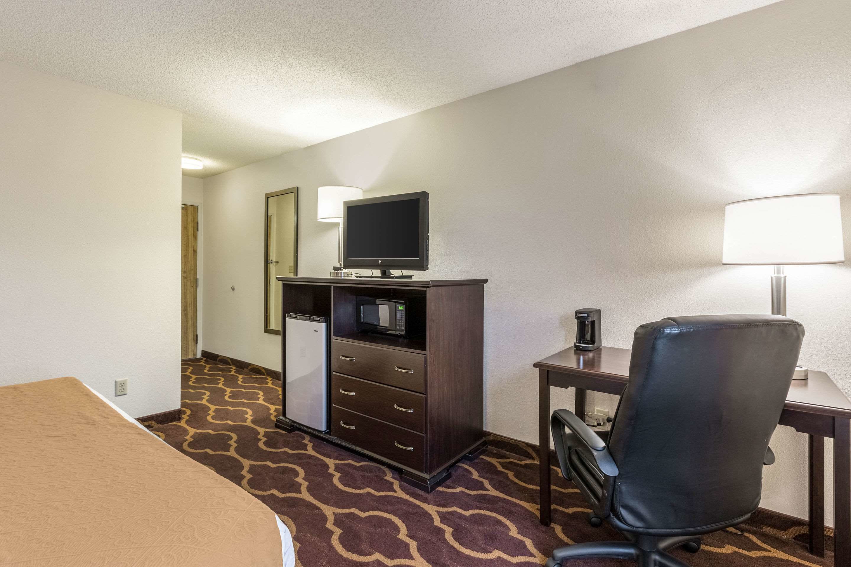 A hotel room with a bed, a desk with a lamp, a television, a minibar, and a chair.