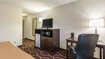 A hotel room with a bed, a desk with a lamp, a television, a minibar, and a chair.