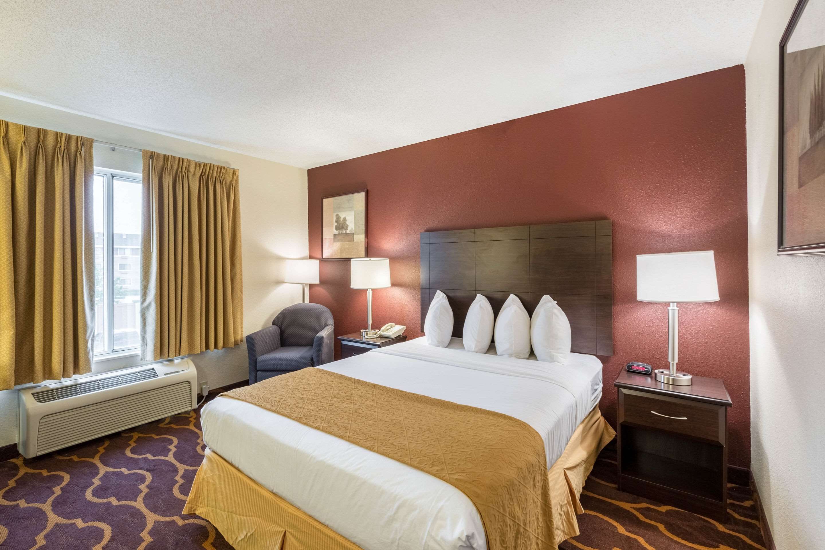 A hotel room with a large bed, two bedside tables with lamps, a chair, a window with curtains, and a wall-mounted picture.