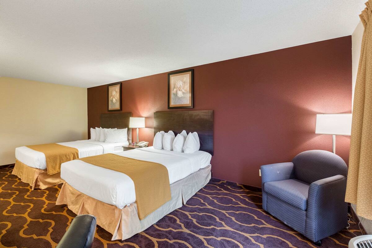 Quarto de hotel com duas camas, uma cadeira, uma escrivaninha com abajur e um quadro na parede.
