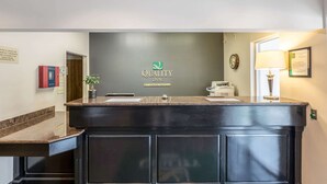 Een receptiebalie met een donker houten afwerking, een logo voor 'Quality Inn' by Choice Hotels, een klok en een ingelijst certificaat aan de muur.