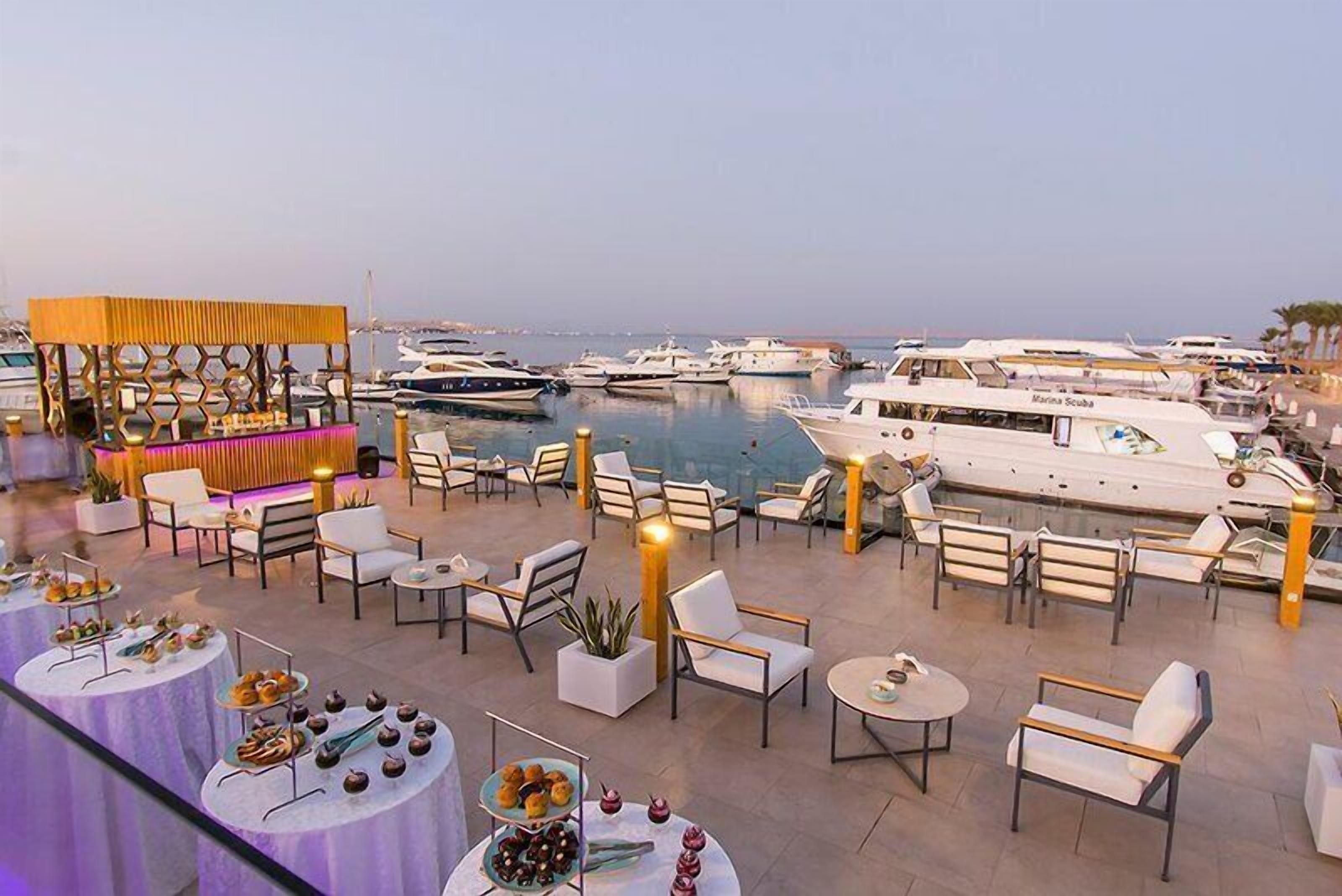 A marina with boats docked, outdoor seating with tables and chairs, and a bar area with a decorative structure.