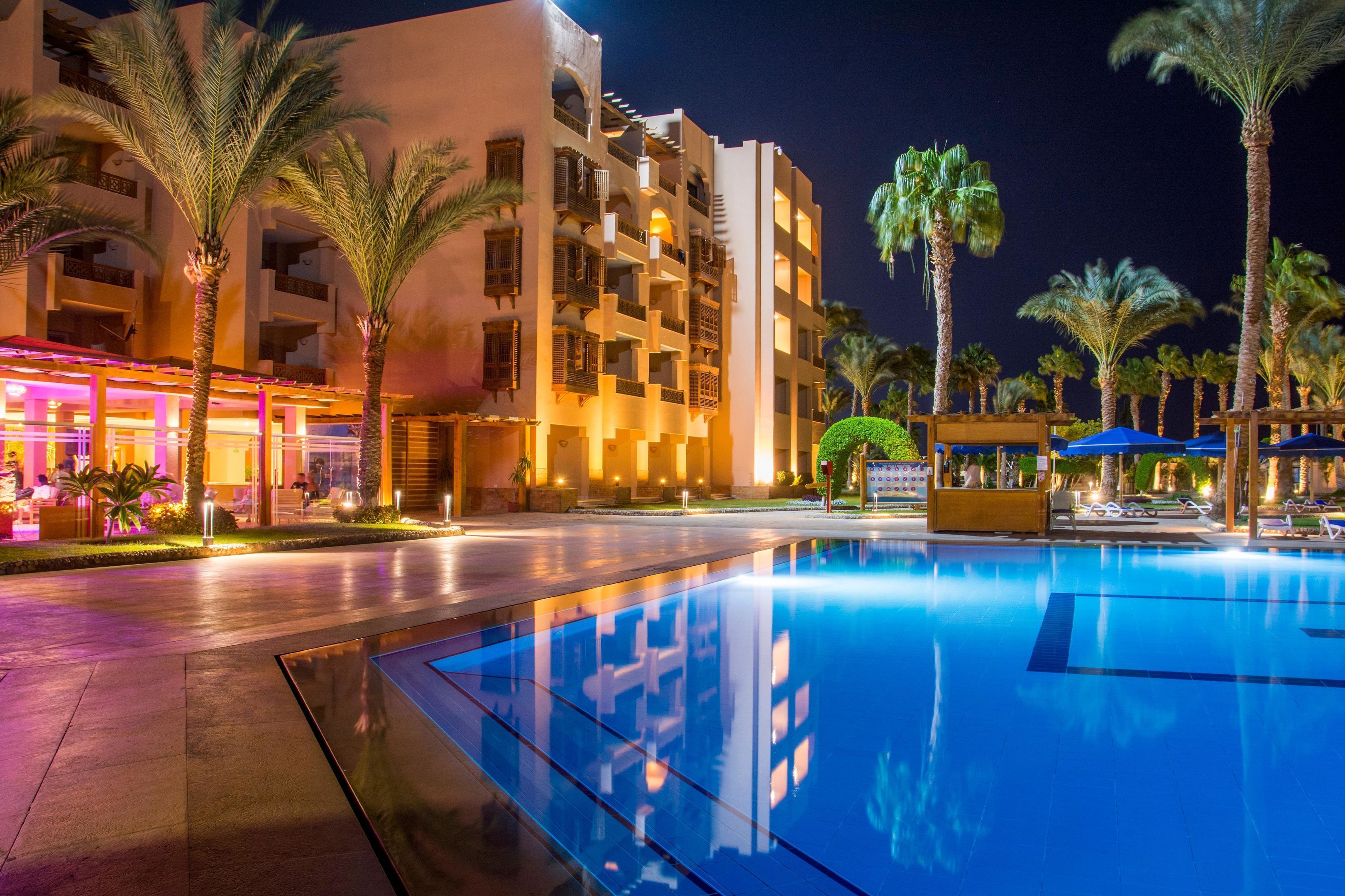 A swimming pool with a clear blue water surface, surrounded by palm trees and a multi-story building with balconies.